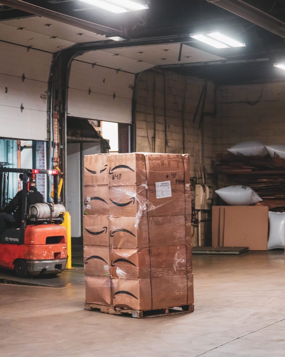 Tall shrink-wrapped pallet of Amazon-branded aid boxes in a warehouse bay, with a forklift near the open dock door