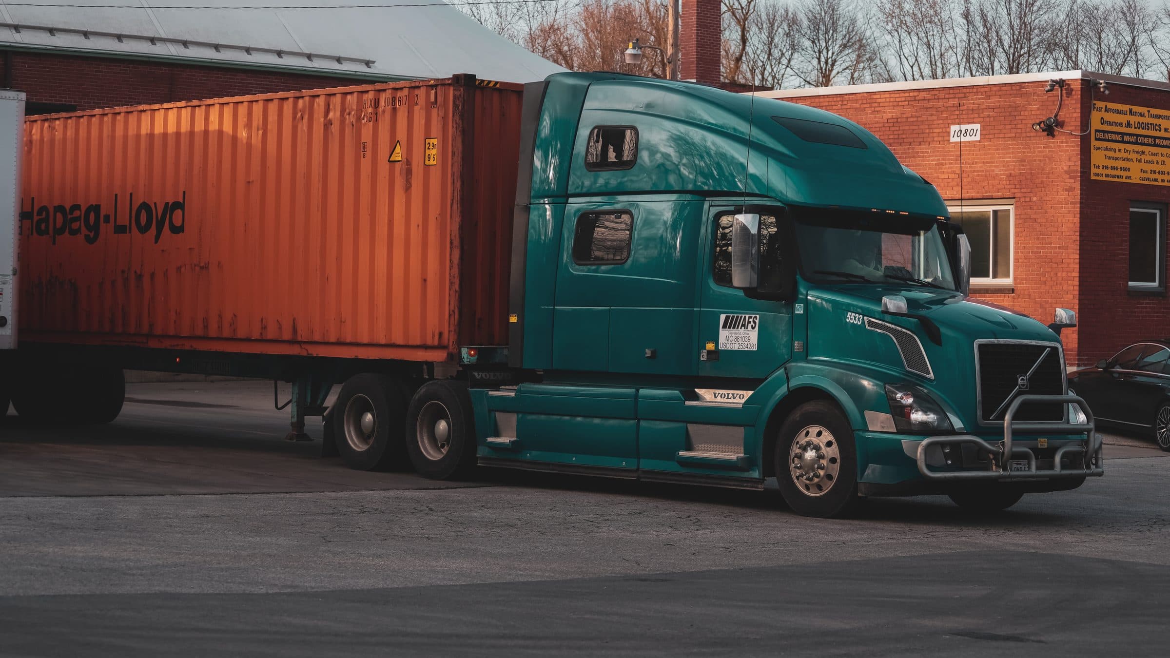 Teal Volvo semi-truck hauling an orange Hapag-Lloyd shipping container at a Cleveland-area warehouse loading dock