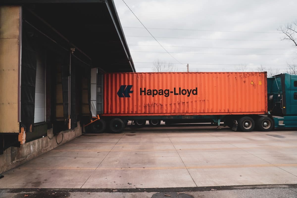 Orange Hapag-Lloyd shipping container on a teal truck backed into a warehouse loading dock